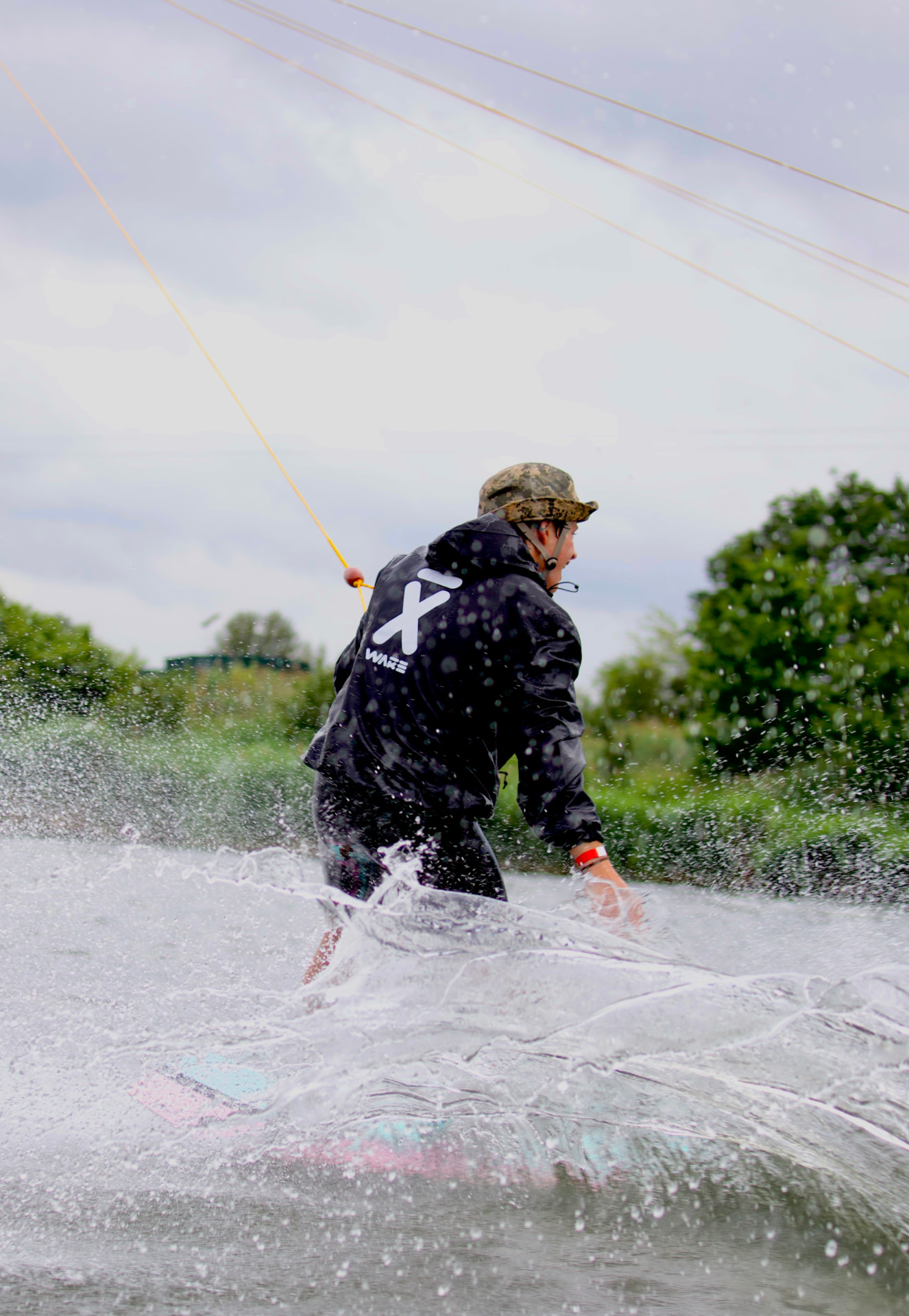 Wakeboarder in black WAKE jacket carving through spray at a cable park